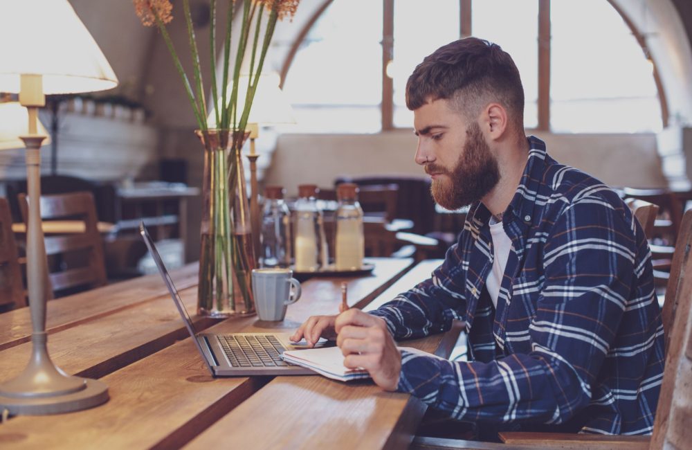 Bărbat în fața unui laptop care scrie pe un carnet de notițe